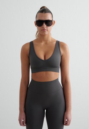 Mujer de pie, mirando hacia adelante, con un sujetador deportivo negro, leggings de talle alto y grandes gafas de sol negras, sobre un fondo blanco liso.