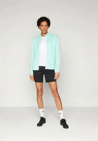 Gaiši zila zip-up jaka, balts T-krekls, melni šorti, balti šķīvji, melnas kurpes. Modelim ir īsi, cirtaini mati, stāvot uz vienkārša fona.