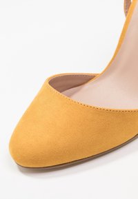 Żółty zamszowy but na wysokim obcasie z ostrym noskiem i otwartym tyłem. Gładka tekstura z subtelnym przeszyciem wzdłuż brzegów.