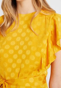 Gul klänning med korta volangärmar, med en texturerad tyg och ett stort cirkulärt prickmönster. Inkluderar ett matchande knytsärmsbälte.