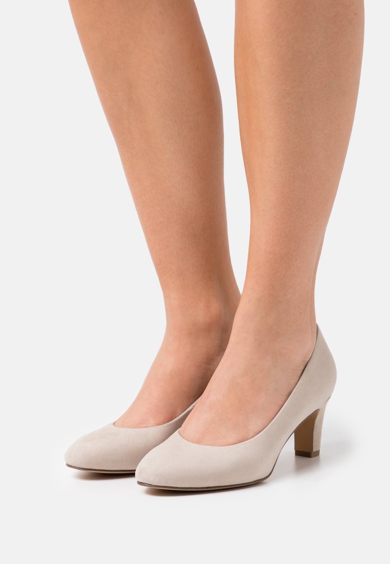 Jambes inférieures d'une femme portant des chaussures en daim beige clair à talons moyens et bouts arrondis sur fond blanc uni.