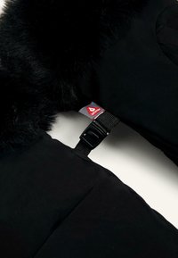Crna jakna od puffer materijala s umjetnim krznom oko vrata, koja ima teksturiranu površinu, remen s kopčanjem i crveno-sivi oznaku brenda.