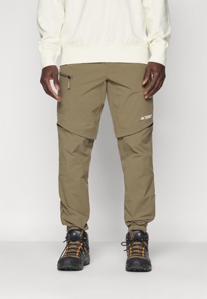 Person trägt olivgrüne Outdoor-Zip-Off-Hose, beiges Sweatshirt und schwarze Wanderschuhe mit orangen Schnürsenkeln, steht vor einem einfarbigen Hintergrund.