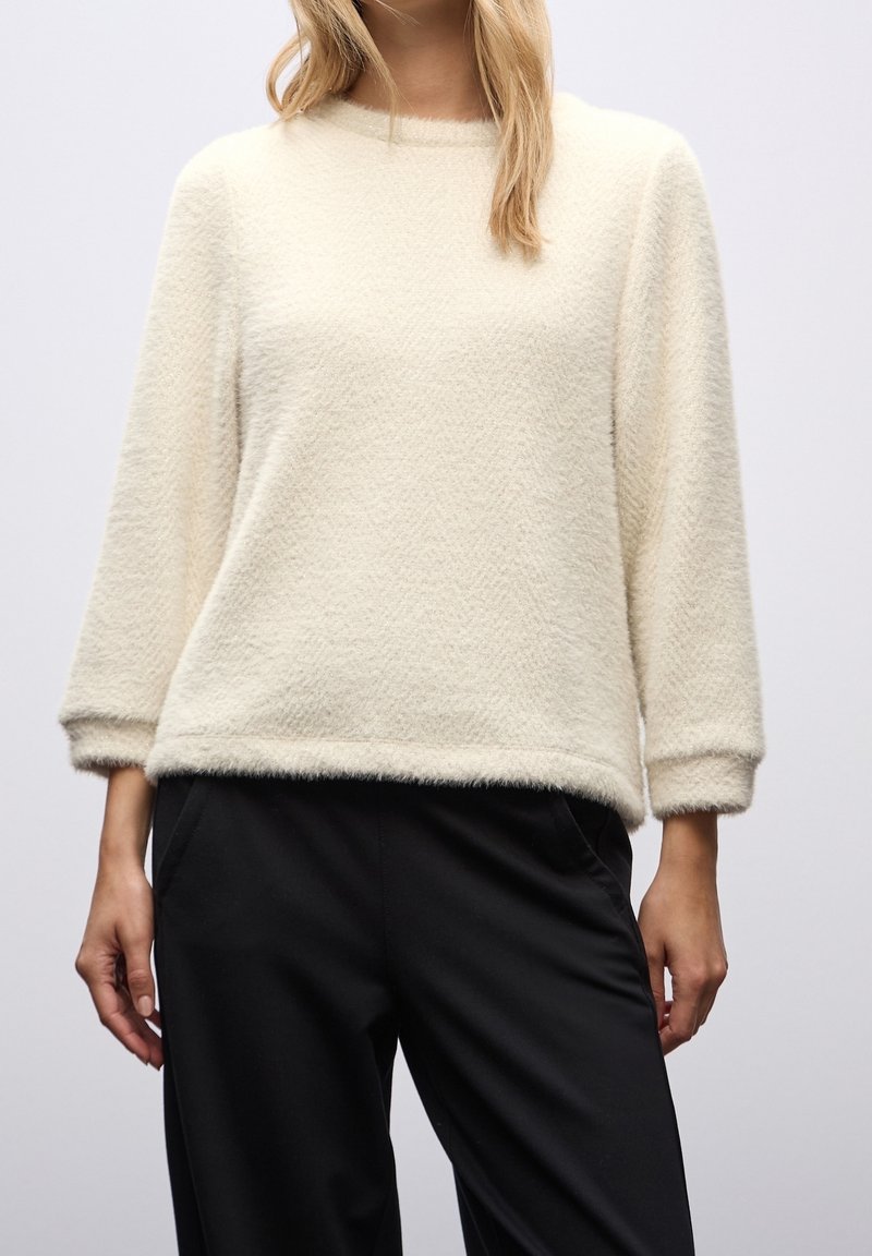 Femme portant un pull doux et pelucheux couleur crème avec des manches trois-quarts et un pantalon noir ample, debout devant un fond uni.