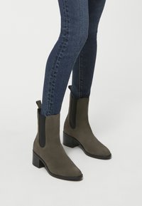 Zielone zamszowe botki za kostkę z elastycznymi panelami bocznymi, kwadratowym noskiem i obcasem w kształcie bloku, zestawione z ciemnymi dżinsami.