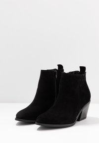 Botines de ante negros con punta afilada, detalle de cremallera lateral y tacón bloque de altura media. Textura suave con un acabado mate.