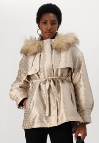 Zlata vzorjena jakna z kapuco obrobljeno z krznom, pasom in stranskimi žepi. Narejena iz sijočega materiala z embosiranim dizajnom.