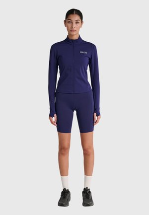 Frau steht mit Blick nach vorne, trägt eine marineblaue langärmelige Jacke mit Reißverschluss, passende Radhosen, weiße Socken und schwarze Sneakers.