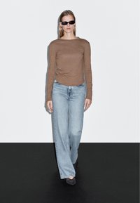 Langarmshirt in Tan, kombiniert mit hellblauen, weiten Jeans. Die Schuhe sind schwarze High Heels. Der Hintergrund ist schlicht weiß mit einem schwarzen Boden.