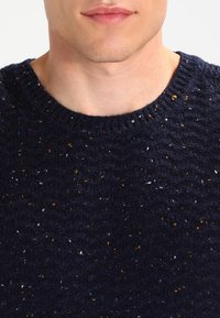 Navy knit sweater featuring a textured pattern, ribbed round neckline, and multicolored speckles in orange, blue, and white throughout.