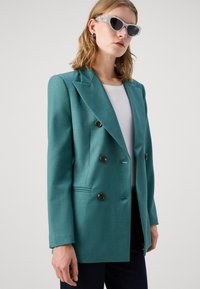 Teal kaksirivinen bleiseri, joka on valmistettu teksturoidusta kankaasta ja jossa on ruskeat napit, huippukaulukset ja sivutaskut. Puettuna valkoisen yläosaan.