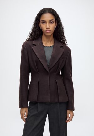 Femme aux longs cheveux bouclés portant un blazer marron foncé cintré sur un haut gris et un pantalon à carreaux foncé, debout devant un fond blanc uni.