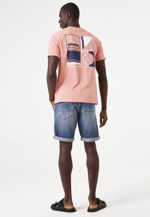 Korte mouwen roze katoenen t-shirt met marineblauw grafisch ontwerp en patronen op de achterkant, gecombineerd met opgerolde denim shorts en zwarte sandalen.