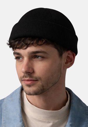 Junger Mann mit kurzen, lockigen Haaren, der eine schwarze Strickmütze, eine helle Jeansjacke und ein cremefarbenes Hemd trägt und leicht nach links schaut.