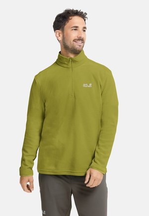 Uomo che indossa una giacca in pile verde con zip e pantaloni scuri, guarda di lato con un leggero sorriso su uno sfondo chiaro.