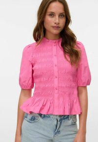 Rosa skjorta med puffärmar, smockad midja, volangkant och slät bomullstextur, kombinerad med ljusblå jeans.