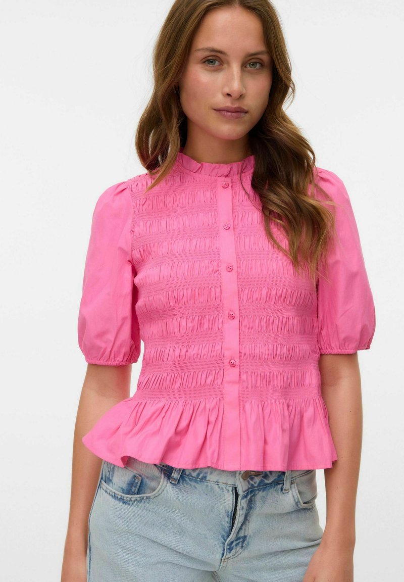 Rosa skjorta med puffärmar, smockad midja, volangkant och slät bomullstextur, kombinerad med ljusblå jeans.