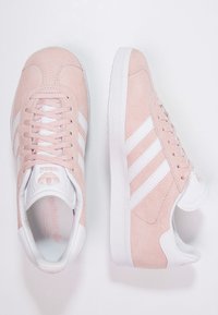 Růžové semišové tenisky s bílým designem tří pruhů, bílou gumovou podrážkou a kulatou špičkou. Obsahují odpovídající tkaničky a polstrovaný límec pro pohodlí.