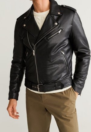 Uomo che indossa una giacca in pelle nera da motociclista con tasche con zip e vita con cintura sopra un maglione bianco e pantaloni marroni.