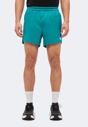 Mann trägt türkisfarbene Laufshorts, weiße Sportsocken und schwarze Sportschuhe, steht mit auseinander stehenden Füßen vor einem schlichten Hintergrund.
