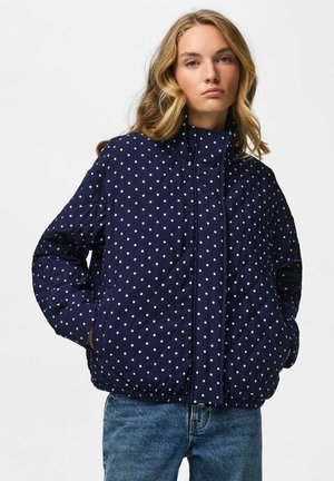 Ung kvinde med blondt hår har en mørkeblå quiltet jakke med hvide polkaprikker og blå jeans på, står med hænderne i lommerne.