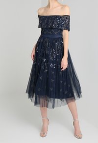 Námořnická modrá šaty s odhalenými rameny a květinovými flitrovými zdobeními. Sukně z tylu dosahuje do poloviny lýtek. Stříbrn�é sandály na podpatku doplňují outfit.
