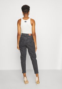 Kvinna med kort lockigt hår iförd en vit ärmlös topp, hög midja mörkgrå jeans och nakna högklackade sandaler, vänd bort.