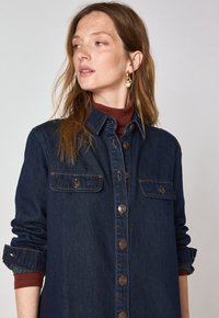 Chemise en denim bleu foncé avec deux poches avant, fermeture par boutons et manches retroussées. Équipements en ton doré et texture douce.