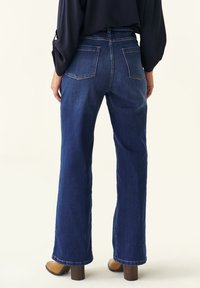 Jeans de pierna ancha en denim azul oscuro con una textura suave, cintura alta y costuras contrastantes. Cuatro bolsillos visibles desde la vista trasera.