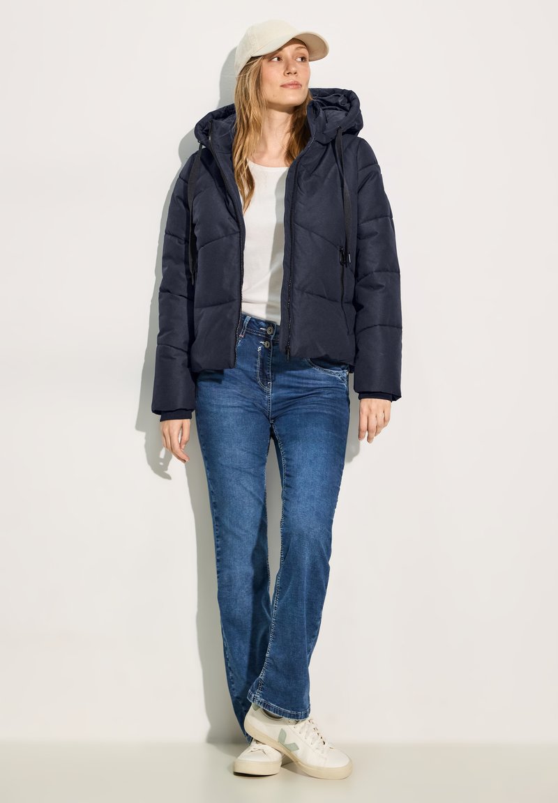 Veste matelassée navy, avec une capuche, une fermeture éclair à l'avant et un design matelassé. Associée à un jean bleu évasé et des baskets claires.