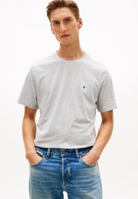 Νέος άνδρας που φοράει ανοιχτό γκρι κοντομάνικο μπλουζάκι και μπλε τζιν, με τα χέρια στις τσέπες, στέκεται μπροστά σε ένα απλό λευκό φόντο.