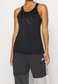 Débardeur de sport noir sans manches avec un logo triangulaire brillant. Le tissu est lisse et ajusté, complété par des bretelles blanches.