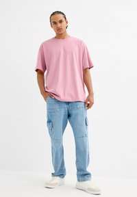 Joven con el cabello trenzado que lleva una camiseta rosa suelta, pantalones cargo azul claro y zapatillas blancas, de pie con una mano en el bolsillo.