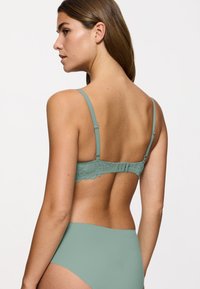 Reggiseno verde in pizzo con spalline sottili e regolabili e bordo ondulato. Vita in tessuto liscio. La vista posteriore mostra la chiusura a gancio e occhiello.