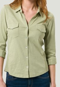 Camicia button-up verde chiaro con due tasche sul petto, maniche arrotolabili e un tessuto testurizzato. Presenta bottoni bianchi e una silhouette dalla vestibilità rilassata.