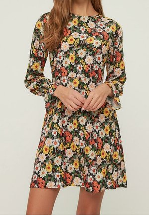 Kvinde iført en kjole med lange ærmer og knælængde med et farverigt blomsterprint på en mørk baggrund.