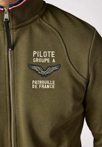 Veste zippée vert olive avec un col montant. Comprend un texte brodé en blanc et bleu et un motif d'aile. Fabriquée en tissu lisse.