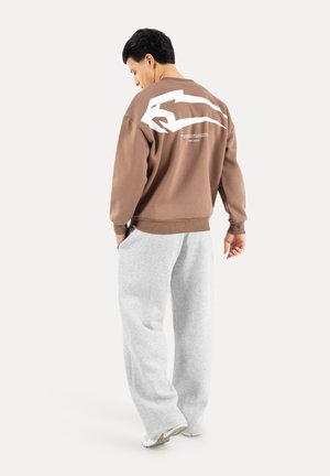 Mand iført brun sweatshirt med hvid grafik på ryggen, lysegrå joggingbukser og hvide sneakers, står og kigger ned.