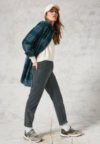 Koszula w kratę w ciemnozielonym i niebieskim kolorze, biała bluza, szare spodnie i zielone buty sportowe z jasnymi i ciemnymi akcentami.