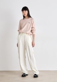 Suéter rosa claro com um logotipo verde, combinado com calças brancas largas com cordão e bolsos laterais, complementadas por ténis pretos.