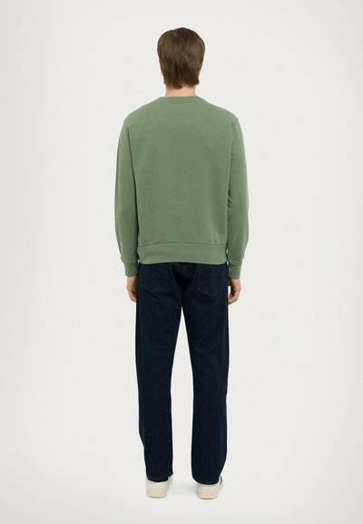 Felpa verde con vestibilità rilassata e polsini a costine, abbinata a jeans blu scuro a gamba dritta e sneakers di colore chiaro.