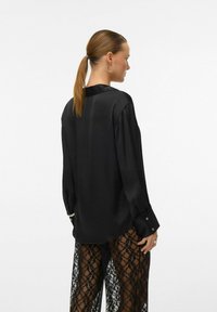 Crna bluzica dugih rukava s sjajnim završetkom, s ovratnikom i dugmadima na manžetama, u kombinaciji s prozirnim crnim čipkastim hlačama.