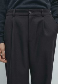 Černé kalhoty na míru s hladkým předním dílem, sklady, poutky na pásek a zapínáním na jeden knoflík. Viditelná hladká textura a strukturovaný střih.