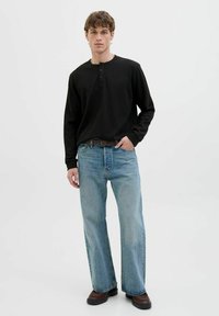 Shirt henley noir à manches longues avec trois boutons, associé à un jean large bleu clair et des bottes marron, sur fond neutre.