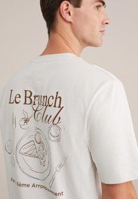 Balts īsām piedurknēm T-krekls ar brūnu ilustrētu dizainu, kas attēlo ēdienu un galda piederumus, un uzrakstu "Le Brunch Club" aizmugurē.
