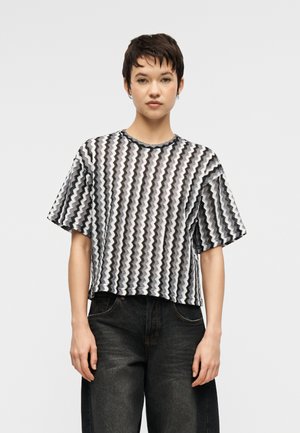 Femme aux cheveux courts portant un haut à manches courtes ample à motifs zigzag noir et blanc et un jean noir délavé, debout devant un fond uni.