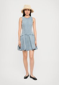 Woman wearing a sleeveless light blue denim dress, straw hat with frayed edges, and black flats, standing against a plain white background.