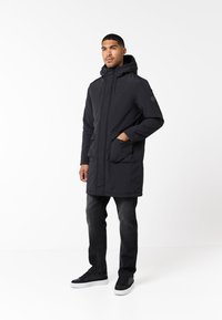 Parka negra con capucha, de manga larga, cierre frontal, dos bolsillos en la parte delantera y un acabado de tela texturizada. Combinada con vaqueros oscuros y zapatillas negras.