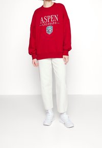 Sudadera roja con el texto "ASPEN COLORADO" y un parche de logo. Presenta un corte relajado, puños acanalados, combinada con pantalones blancos y zapatillas.