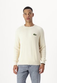 Cremfarbener, gerippter Pullover aus strukturiertem Strickstoff, mit Rundhalsausschnitt und kleinem, grünem, gesticktem Krokodil-Logo auf der Brust.